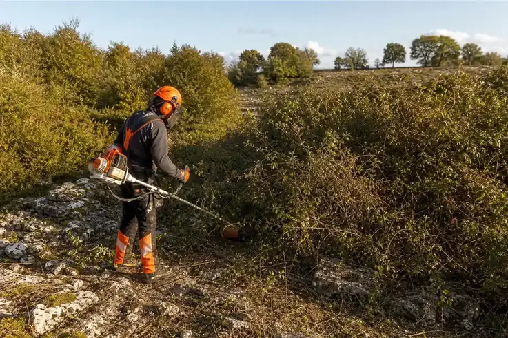 Photo d'un paysagiste en tenue de sécurité débroussaillant à la débroussailleuse à dos un terrain envahi de ronces sur le causse autour de Rodez, lumière de fin de journée, sol calcaire caillouteux visible.