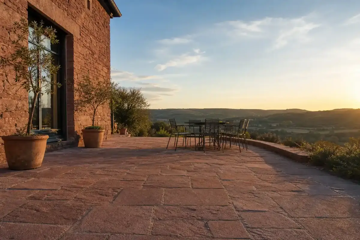 Terrasse en grès rose du Rouergue devant une maison ruthénoise, mobilier de jardin sobre, lumière chaude de fin d'après-midi, causse en arrière-plan, cadrage large au ras du sol montrant les joints et la pente.