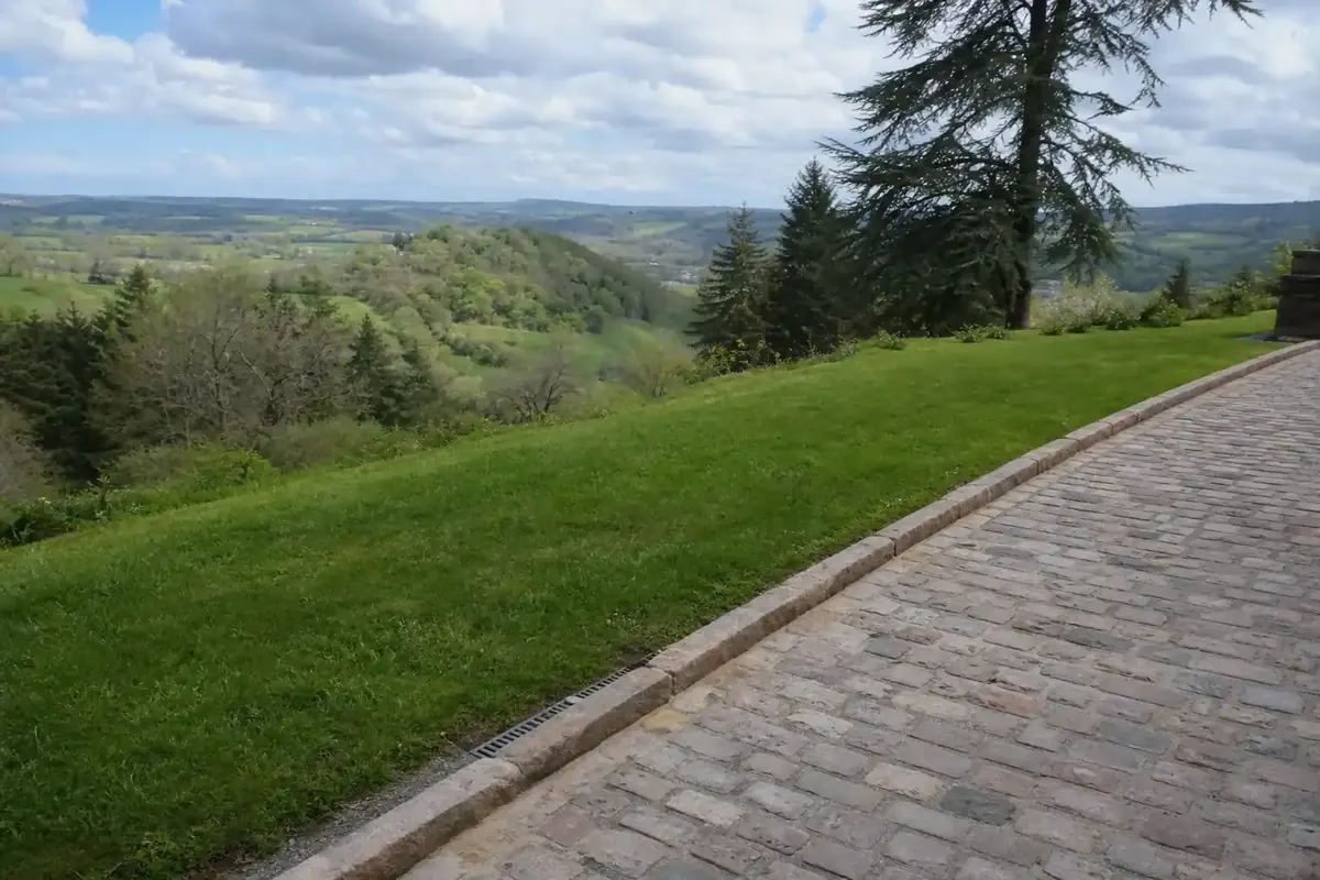 Allée de pavés bordée d'une bordure pierre le long d'une pelouse, terrain légèrement en pente du Ségala, drain visible en rive, ciel de l'Aveyron.