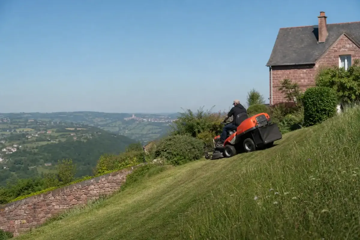 Jardinier tondant une pelouse en pente sur un coteau ruthénois avec une tondeuse autoportée, terrain vallonné du pays ruthénois en arrière-plan.