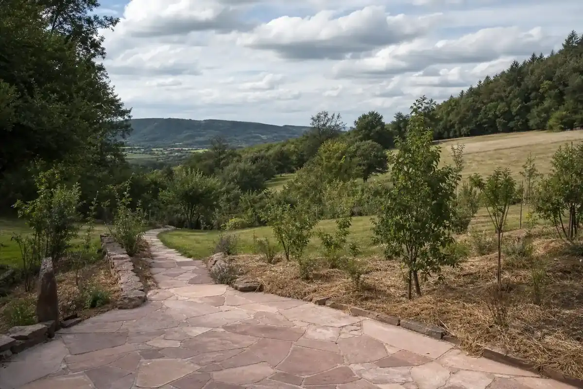 Vue d'une terrasse en pierre locale reliée par une allée à un jardin en pente du pays ruthénois, avec de jeunes arbustes plantés et paillés, sous un ciel du Massif central.