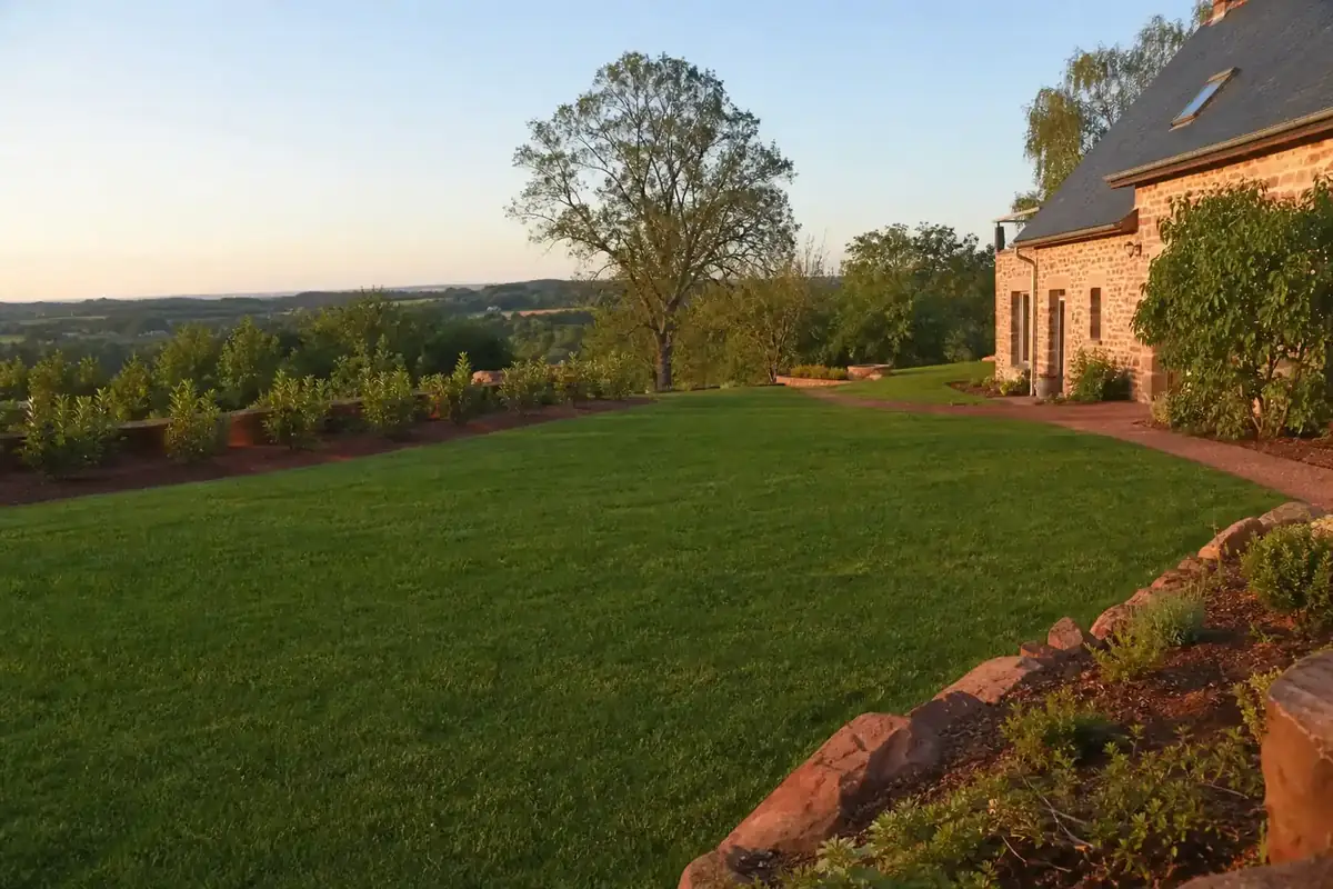 Grand jardin fraîchement engazonné devant une maison en pierre près de Rodez, gazon vert dense bien net, terre rouge du Rougier visible en bordure, lumière douce de fin de journée sur le plateau ruthénois.