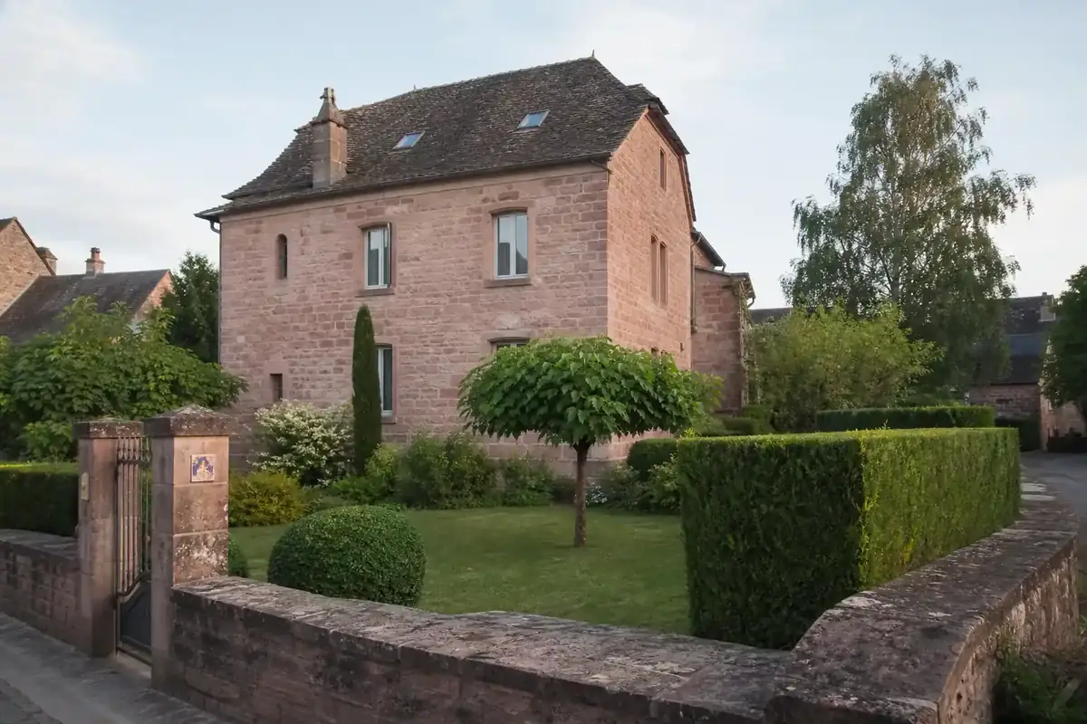 Photo d'un jardin ruthénois soigné devant une maison en pierre, pelouse fraîchement tondue et haies nettes, façade en grès rose du Rouergue en arrière-plan, lumière douce de fin d'après-midi, cadrage large depuis la rue.