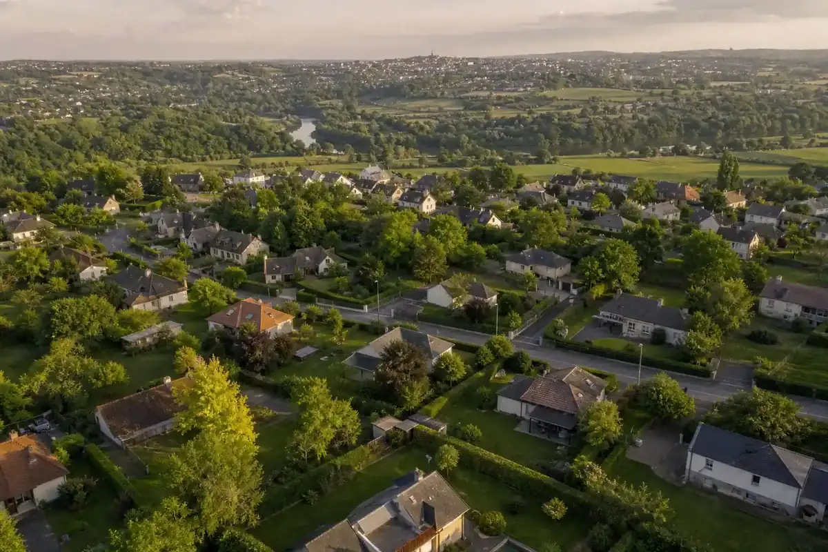 Vue aérienne basse d'un quartier pavillonnaire de la couronne de Rodez : jardins verts contigus, haies taillées, la vallée de l'Aveyron en contrebas du piton, lumière de fin d'après-midi.